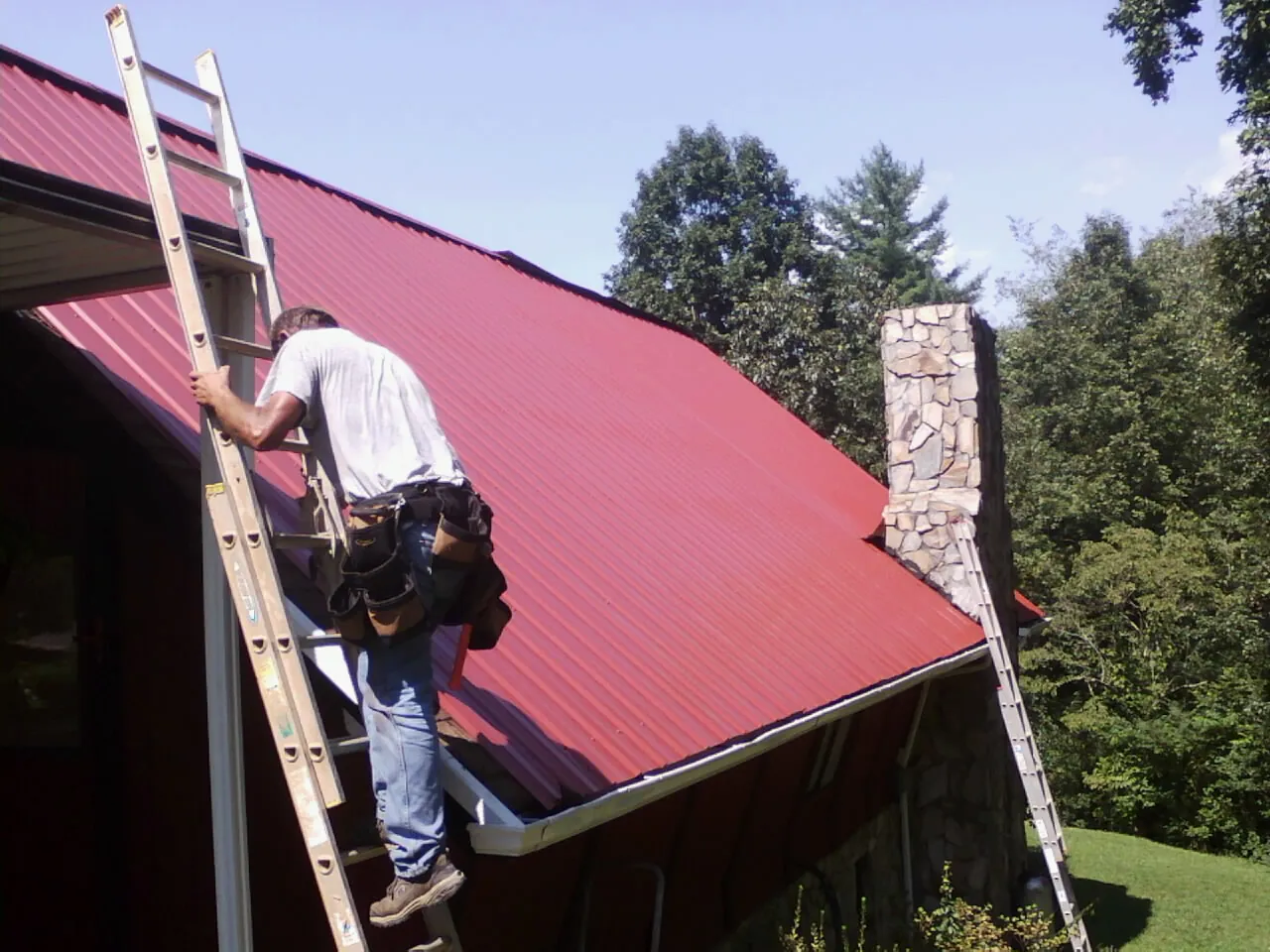 Roofer on a ladder beside a red metal roof and stone chimney — Soffit & Fascia Repair by Roofing Brothers in Boiling Springs